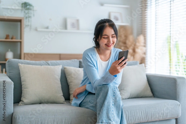 Obraz Smiling pretty young Asian woman sitting on cozy couch, using funny mobile apps in living room. Woman at home, Doing online shopping, Messaging friends, Posting on social media