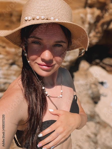 Obraz A cheerful young woman in a straw hat and bikini smiles while taking a selfie at the beach. Surrounded by the coastline and natural beauty, she embodies summer relaxation and enjoyment in Melbourne