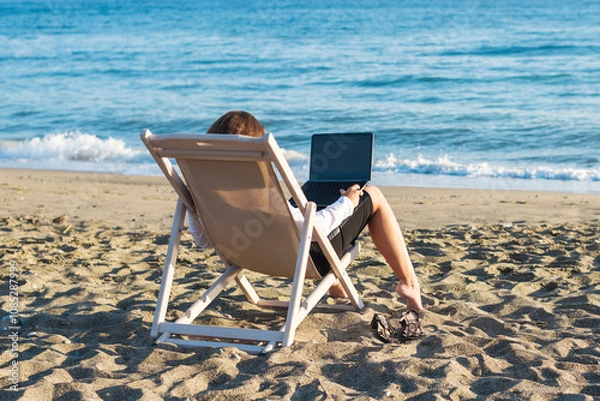 Fototapeta Back view of alone young brunette woman freelancer in office clothes with a laptop looking on the sea sitting on the deck chair on beach. Summer concept