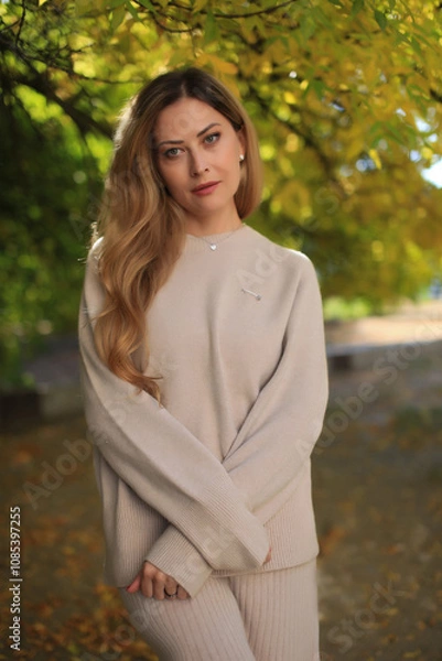 Fototapeta A beautiful fashionable young blonde girl stands in an autumn park on leaves in a gray dress and sneakers
