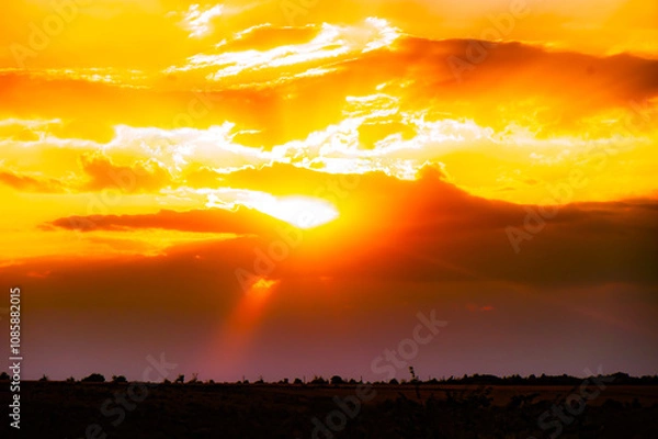 Fototapeta Vibrant Sunset with Sun Rays Breaking Through Clouds. Dramatic sunset with golden rays piercing through vibrant orange clouds, illuminating the rural landscape below.