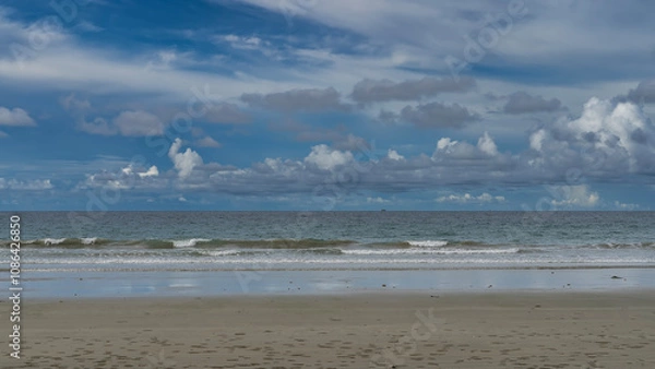 Fototapeta The waves of the endless turquoise ocean foam and spread over the beach. Reflection on wet sand. Sand bubbler crabs burrows are visible. A tiny silhouette of a ship on the horizon. Blue sky, clouds.