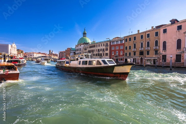 Fototapeta A large public transportation vaporetto near Ferrovia stop, moves along the Grand Canal in Venice. Other smaller boats are visible, along with the historic architecture of the city. Vibrant blue sky.