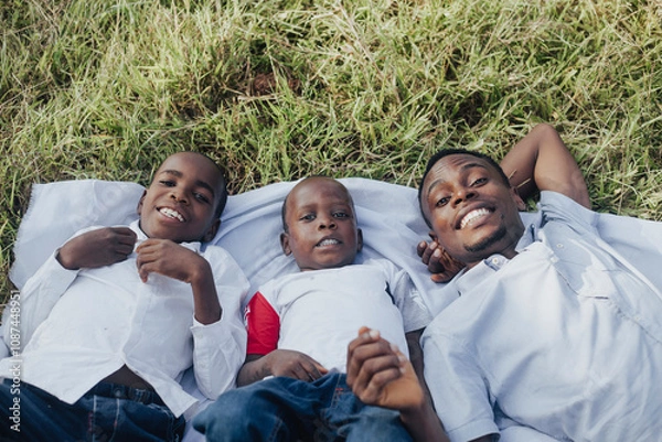 Fototapeta A happy family of three, wearing white shirts, lies on the grass enjoying a sunny day outdoors. They are smiling joyfully, showcasing warmth and togetherness.