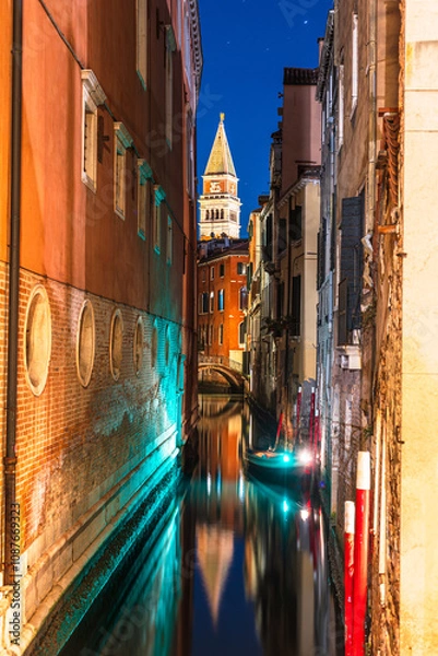 Fototapeta A narrow canal in Venice, Italy, at night, with a gondola passing under a bridge and a tall bell tower of St Mark's Campanile visible in the distance.