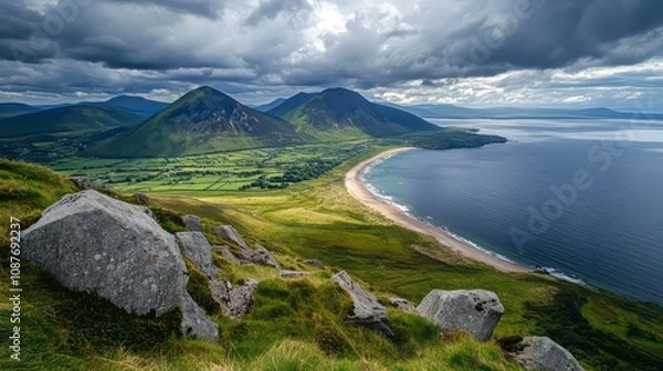 Fototapeta A scenic view of a coastal landscape with green hills and a long, sandy beach. The sky is cloudy, with a bright sun shining through the clouds. There are large boulders in the foreground.