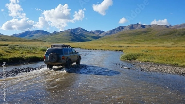 Fototapeta An off-road vehicle traversing a river in a mountainous landscape under a clear blue sky.