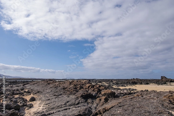 Obraz White sand beach with solidified volcanic lava flows, black rocks. Mountains in the background and sky with big white clouds. Caletón Blanco, Lanzarote, Canary Islands, Spain