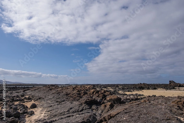 Fototapeta White sand beach with solidified volcanic lava flows, black rocks. Mountains in the background and sky with big white clouds. Caletón Blanco, Lanzarote, Canary Islands, Spain