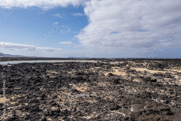 Fototapeta White sand beach with solidified volcanic lava flows, black rocks. Mountains in the background and sky with big white clouds. Caletón Blanco, Lanzarote, Canary Islands, Spain