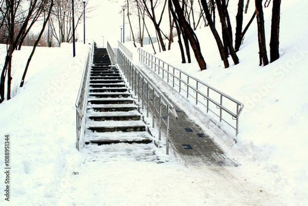 Obraz A snow-covered staircase with a handrail and ramp for wheelchair. Inclusion and winter accessibility in urban environments. Scene featuring a snowy staircase and ramp for people with disabilities