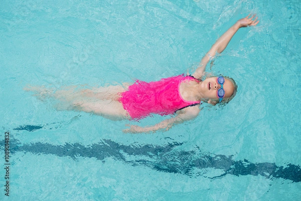 Fototapeta Caucasian girl in pink swimsuit and blue goggles floating on her back in a clear swimming pool