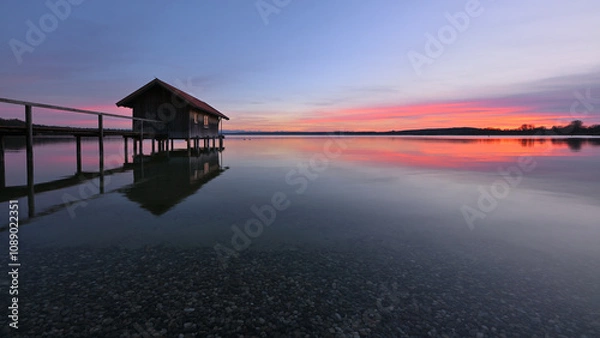 Obraz Ein Bootshaus steht einsam im absolut stillen und klaren Wasser eines See. Ein schönes Abendrot zeichnet sich am Himmel ab, das sich im See Spiegelt.