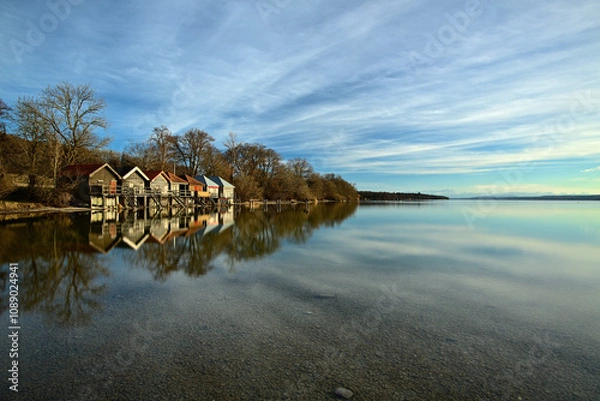 Obraz Bunte Bootshäuser stehen im Frühling an einem idyllischen See, in dem sich die Landschaft spiegelt. Bei leicht bewölkten Himmel kann man bis zu den Alpen sehen.