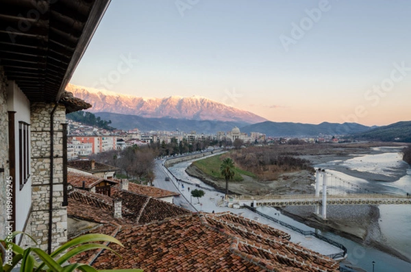 Fototapeta A panoramic view of a Berat with the Osumi river flowing through it. The city is nestled against a backdrop of snow-capped mountains, with a traditional bridge spanning the river. 