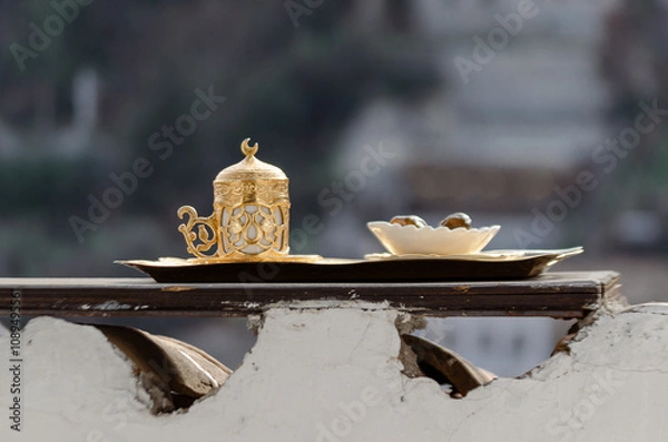 Fototapeta A traditional Turkish coffee set, made of gold and porcelain, rests on a wooden tray. The set is placed on a white wall with a blurred cityscape in the background.