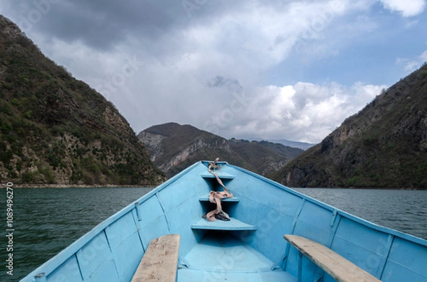 Fototapeta A small, blue boat glides through a serene lake surrounded by towering, lush mountains. The sky is filled with dramatic, fluffy clouds, casting a serene atmosphere.