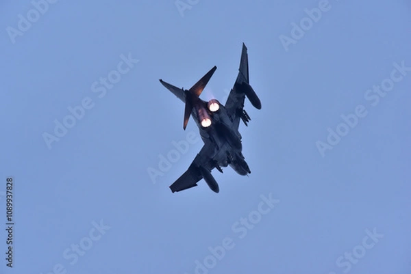 Fototapeta A dynamic photograph of a two engine  fighter jet soaring through the sky with its afterburners glowing brightly. The aircraft is captured from below, highlighting its powerful and aggressive stance 