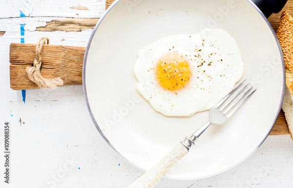 Fototapeta Fried egg with spice and bread slices in white ceramic frying pan on wooden board 