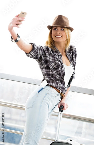 Fototapeta A tourist blonde woman in a striped sweater with a suitcase and passport makes selfie at the station or terminal