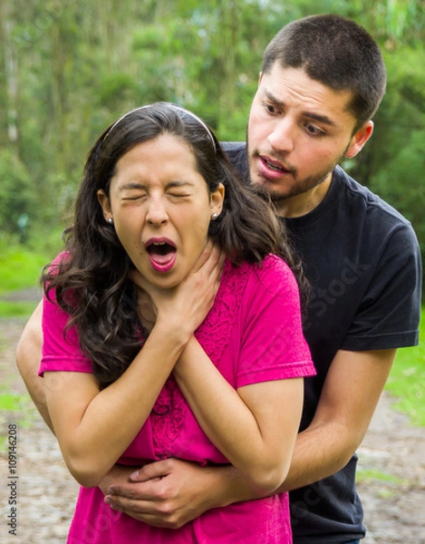 Obraz Young woman choking with man standing behind performing heimlich maneuver, park environment and casual clothes