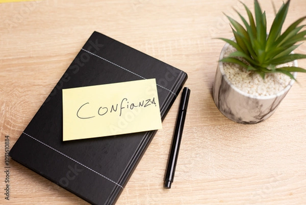 Obraz Notebook with confianza note, pen and plant on wooden desk