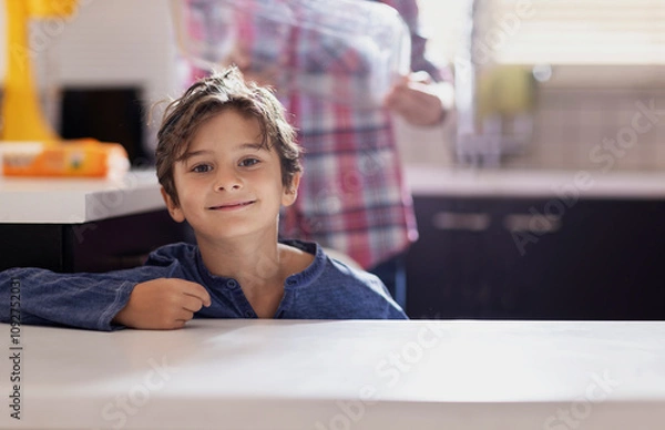 Obraz A young boy with brown hair smiles while leaning on a kitchen counter. The scene shows a cozy kitchen, with a blurred adult in the background preparing breakfast at dawn.