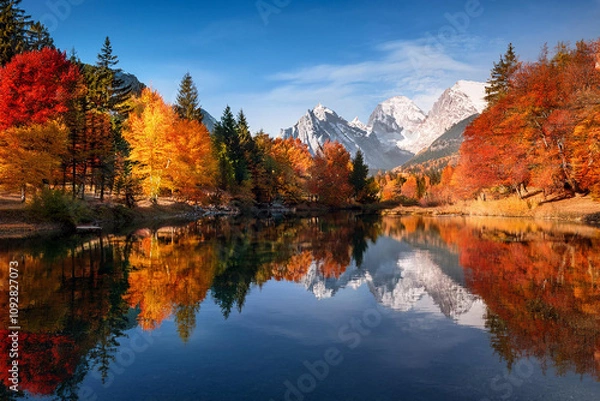 Fototapeta Tranquil lake surrounded by autumn trees and mountains, with vibrant red, orange, and yellow leaves mirrored on the water's surface, ai.