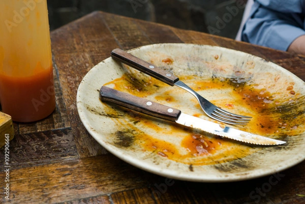 Fototapeta A cream-colored plate with a beautiful pattern shows leftover food, including a knife and fork. Some sauce remains on the plate, indicating the end of a meal