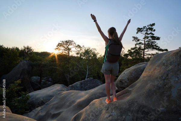 Fototapeta Young woman hiker standing alone with outstretched arms on mountain footpath enjoying view of evening nature on wilderness trail. Active way of life concept