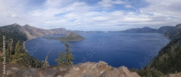Fototapeta Panoramic view of beautiful crystal clear blue lake from high elevation, showing pine trees on the nearby ridge, a dramatic cloudscape over head and in the far distance the other side of the volcanic 