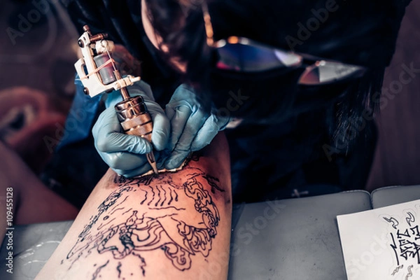 Fototapeta Zbliżenie procesu tatuażu w profesjonalnym salonie. Mistrz wykonuje wzór tatuażu na nodze klienta. Mistrz pracuje w sterylnych niebieskich rękawiczkach.