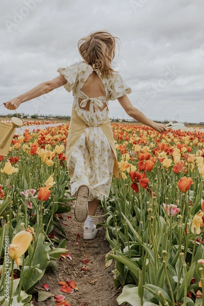 Fototapeta A girl in a yellow dress and apron with a water can in a red and green tulip flower field on a gloomy rainy cloudy sky background happily running around. 

