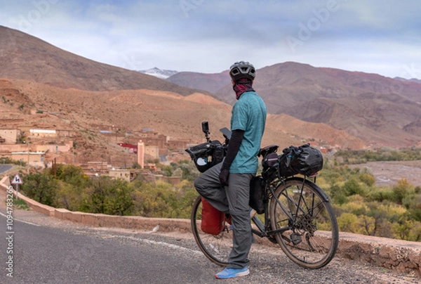 Obraz Bike packer man in Atlas Mountains in Morocco, admires village with mosque, colorful mountains and snow caps. Nomad traveling on his bike.