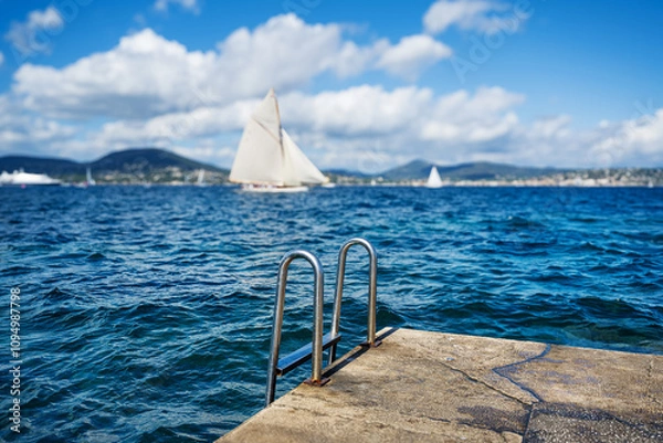 Fototapeta Handrail of a metal chrome ladder leading from a platform into the blue sea in the background sails a historic yacht.