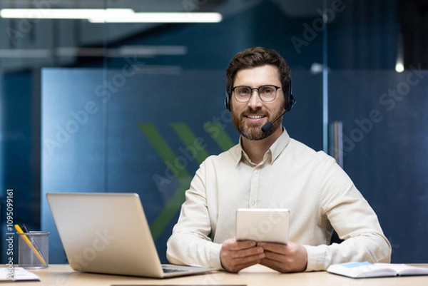 Fototapeta Portrait of a young businessman in a headset sitting at a workplace with a laptop, holding a tablet in his hands and looking at the camera with a smile