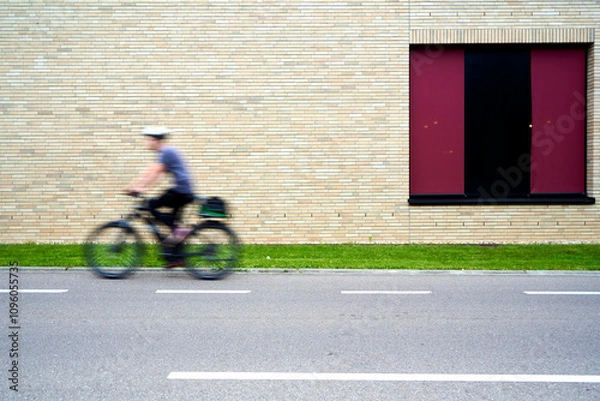 Fototapeta Person riding a bicycle past a brick wall with a large red window, motion blur conveys movement and speed, adding dynamism to the scene.