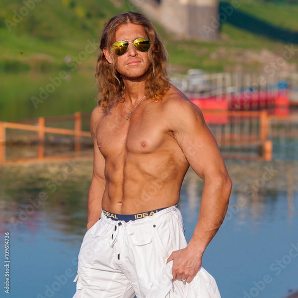 Fototapeta Handsome fitness model shirtless man walks along the beach in the city. Handsome man on the beach in sunglasses