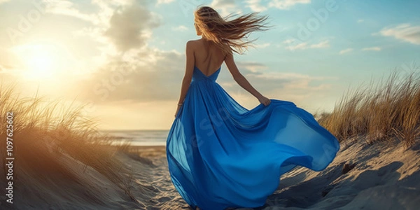 Obraz A woman gracefully adorned in a flowing blue gown stands by a windswept shoreline, her dress dramatically catching the breeze, under a vibrant sky.