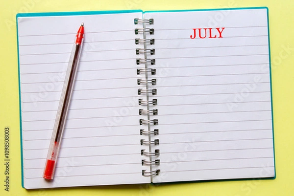 Fototapeta the word JULY written on an open notebook, with a red pen next to , on yellow background. Top view