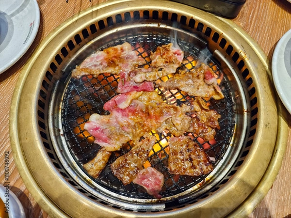 Obraz Top view of sizzling meat slices being grilled on a barbecue grill with glowing charcoal underneath, surrounded by plates on a wooden dining table, showcasing a delicious barbecue experience