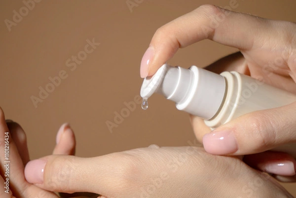 Obraz Drop of serum in close-up drips onto well-groomed female hand from an unmarked beige dispenser.