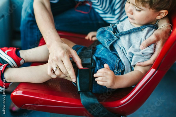 Obraz Mother fastens a seat belt for her son sitting on a bright red chair on a bus. The child looks focused and comfortable, ready for the journey ahead.