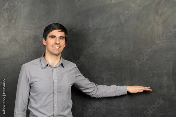 Fototapeta Portrait of young happy smiling teacher man standing near chalkboard background and showing something on it