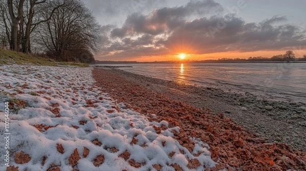 Obraz A serene riverside view at sunset, featuring snow and autumn leaves along the shore.