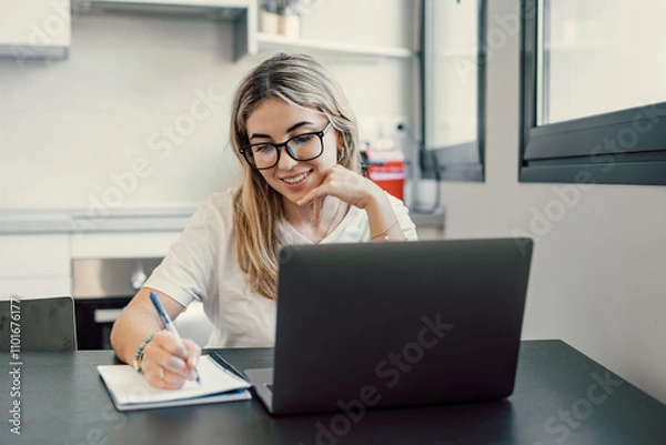 Fototapeta Millennial blonde girl sit at desk in living room study on laptop making notes, concentrated young woman work on computer write in notebook, take online course or training at home, education concept.
