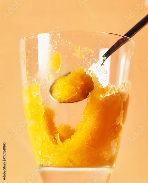 Fototapeta Refreshing Mango Sorbet Served In A Clear Glass With A Spoon. A bright yellow mango sorbet is scooped from a glass, illuminated against a warm orange background