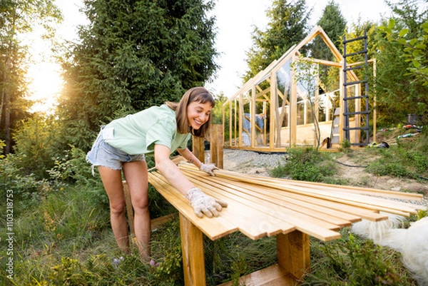 Fototapeta Smiling female carpenter holding wooden planks in front of a partially built greenhouse in her backyard. Engaged in a DIY project, surrounded by nature