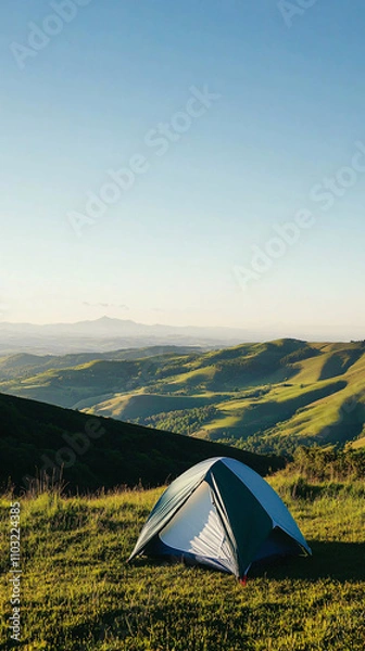 Obraz A small tent sits in a lush green landscape under a clear sky, overlooking rolling hills and distant mountains in a serene and eco-friendly setting