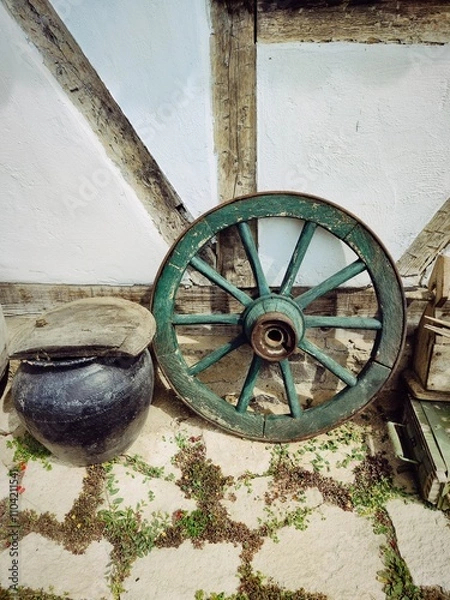 Fototapeta An old retro wheel from a horse carriage paired with a rustic pottery jug, evoking a sense of nostalgia and traditional craftsmanship. This vintage combination captures timeless rural charm.
